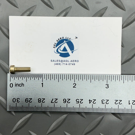 A small metallic pin or screw component placed next to a white card with a blue logo and a metal ruler showing inch measurements, photographed on a gray diamond-plate metal surface.